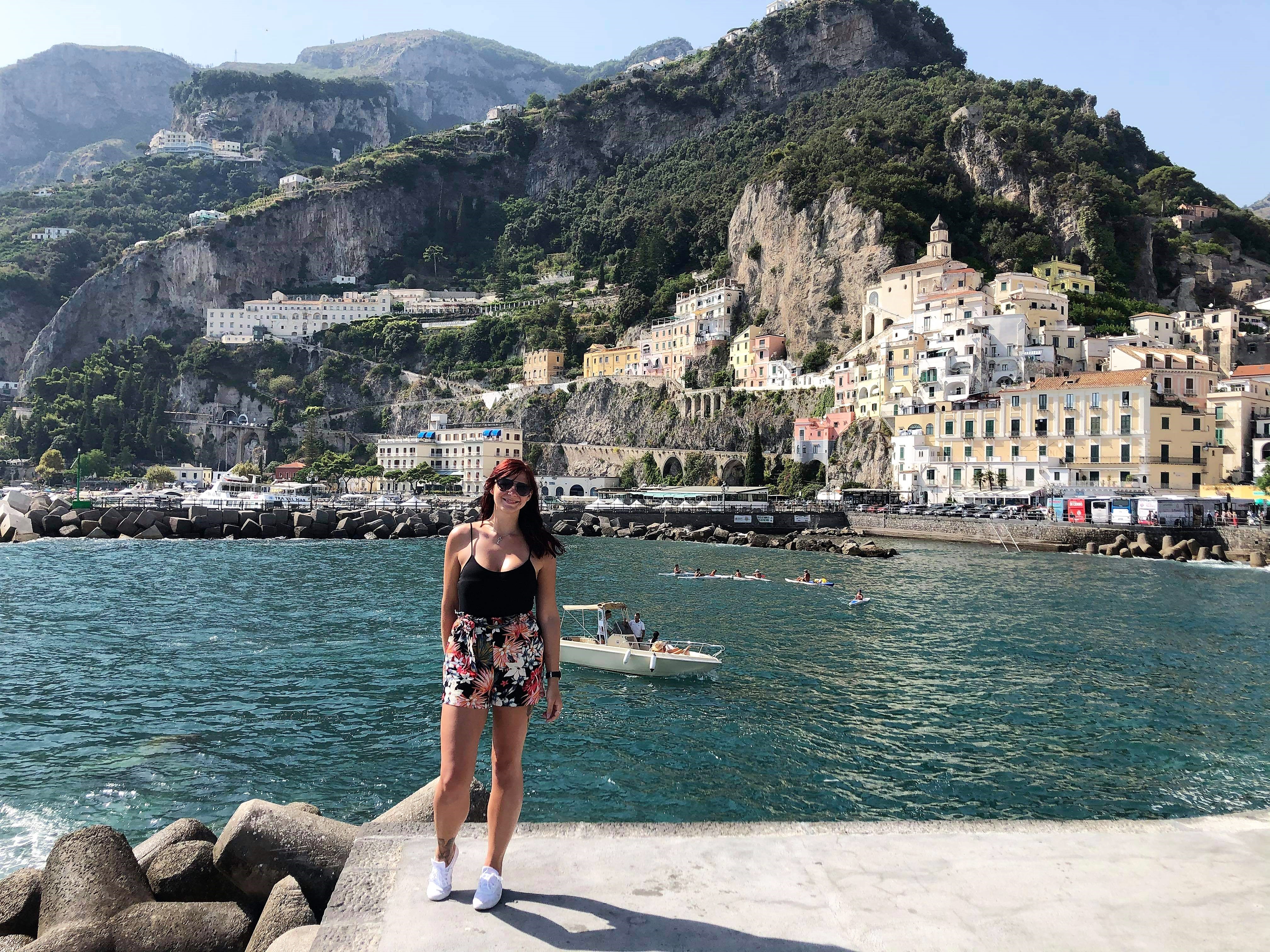 Amalfi Coast Road Trip - female blogger posing for a photo in front of the sea in amalfi with colourful houses and mountains in the background