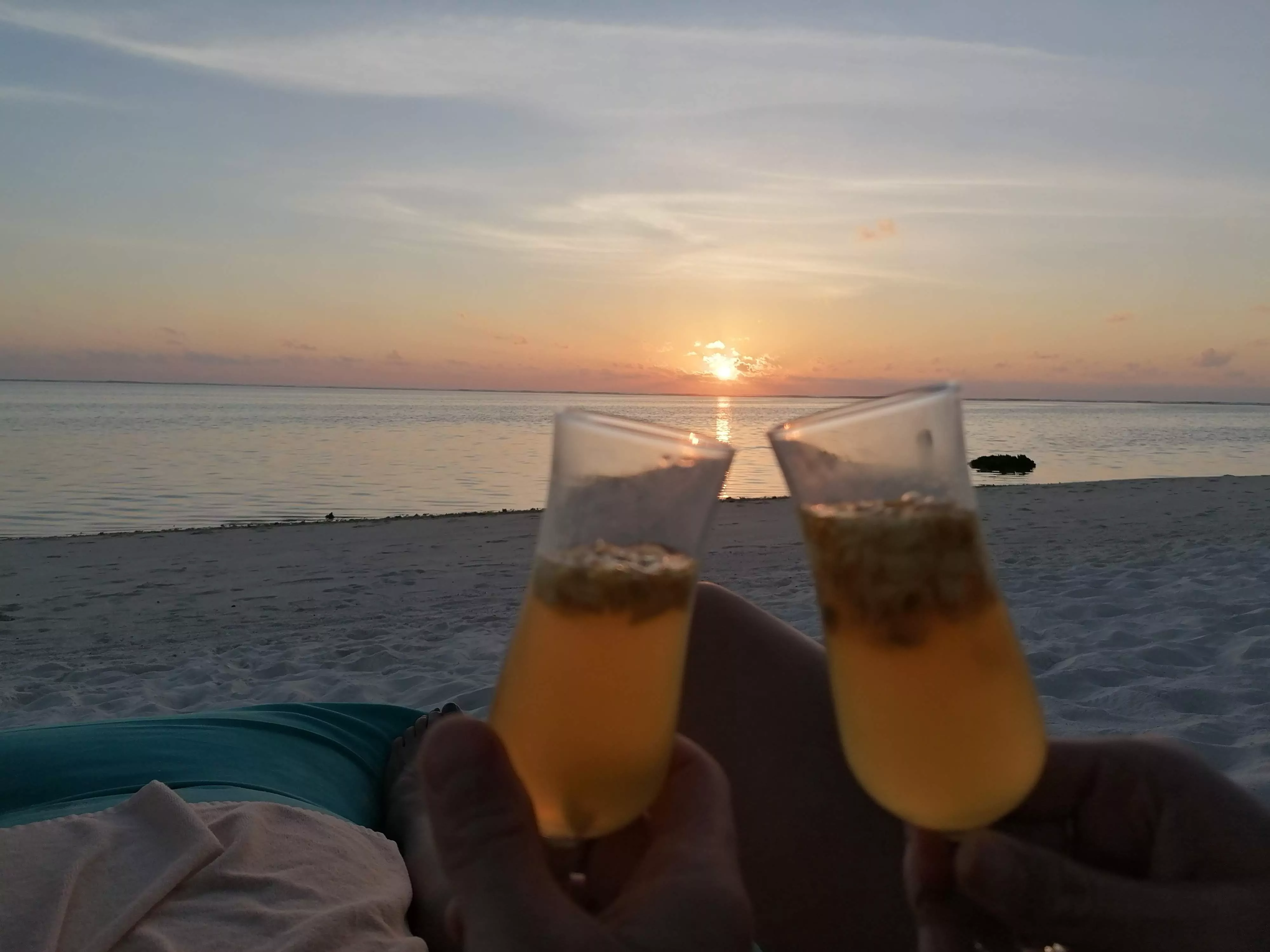 Maldives Travel Guide - two champagne flutes being toasted on the white sands of the maldives as the sky is turning orange with the sunset